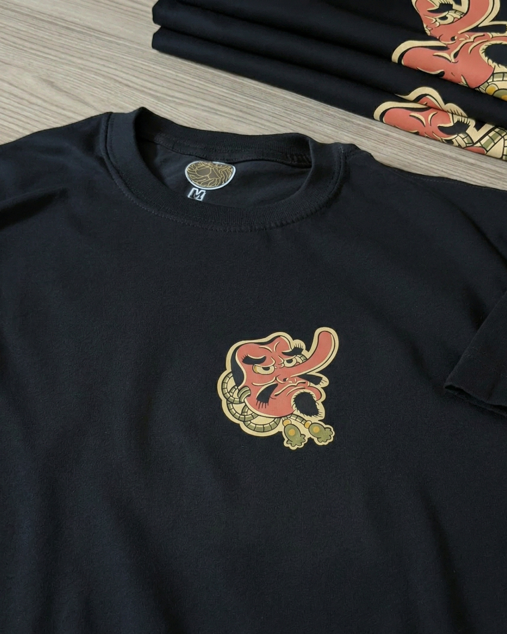 Primer plano detallado del estampado de máscara Tengu sobre una camiseta negra de algodón. Se aprecia la textura de la tela, la etiqueta interna del cuello y la intensidad de los colores rojo y beige
