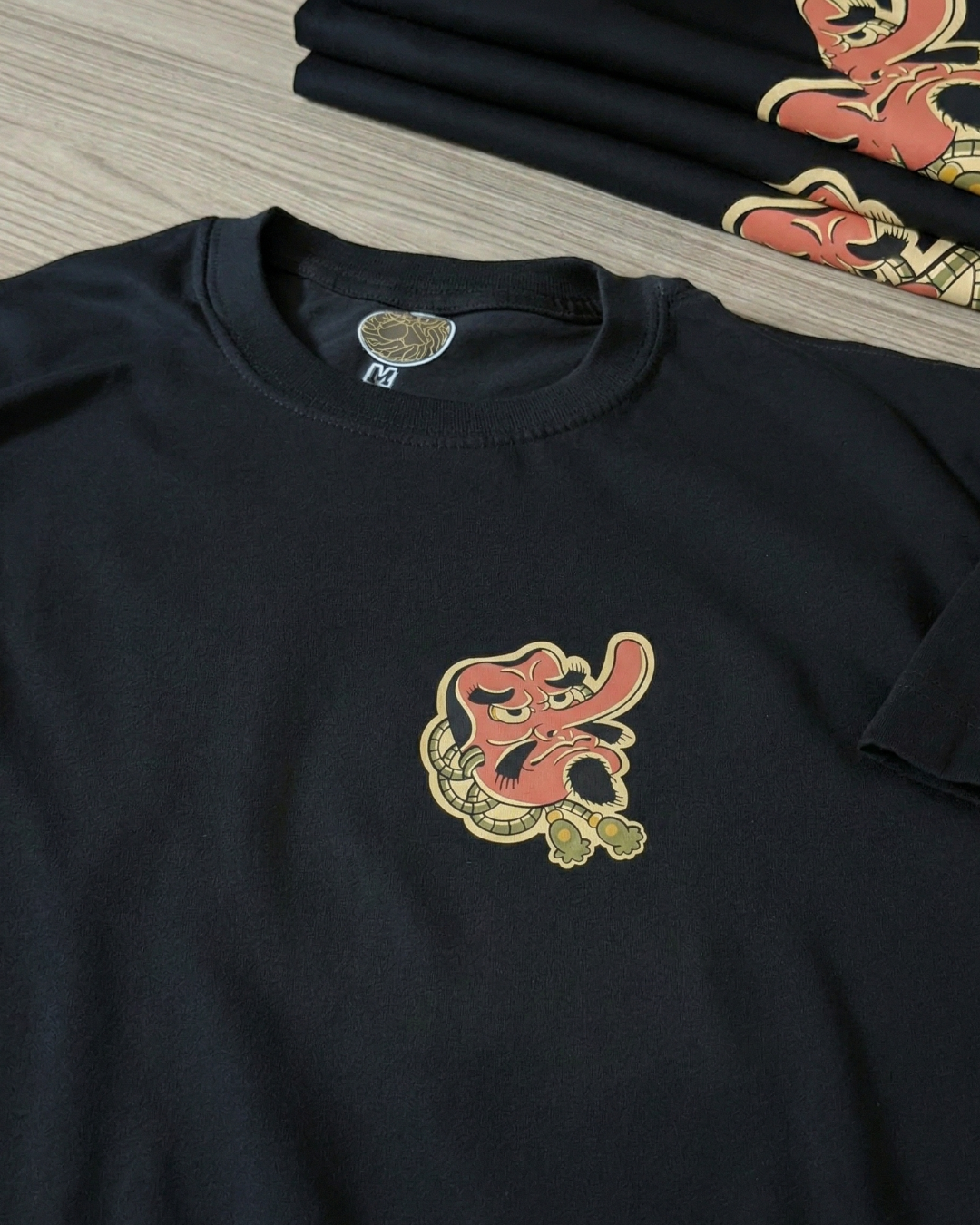 Primer plano detallado del estampado de máscara Tengu sobre una camiseta negra de algodón. Se aprecia la textura de la tela, la etiqueta interna del cuello y la intensidad de los colores rojo y beige