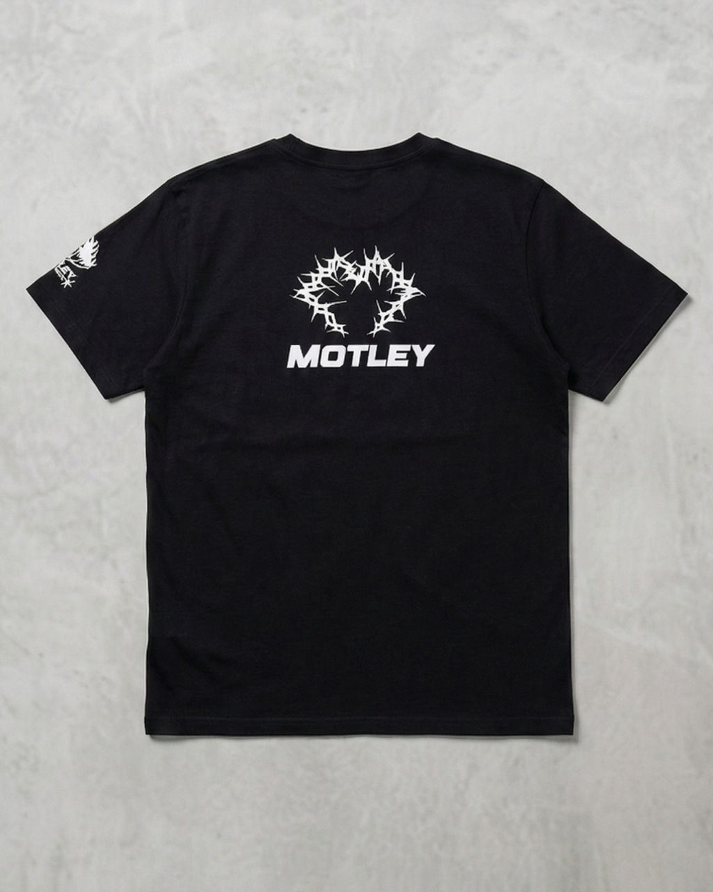 Vista trasera plana de una camiseta negra de manga corta sobre un fondo gris texturizado, con un gráfico central blanco de espinas entrelazadas y la palabra "MOTLEY" debajo en fuente sans-serif blanca, además de un gráfico blanco más pequeño en la manga izquierda.