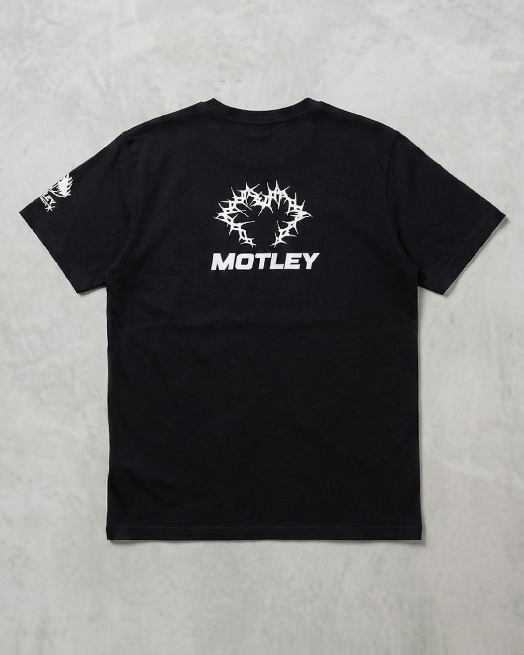 Vista trasera plana de una camiseta negra de manga corta sobre un fondo gris texturizado, con un gráfico central blanco de espinas entrelazadas y la palabra "MOTLEY" debajo en fuente sans-serif blanca, además de un gráfico blanco más pequeño en la manga izquierda.