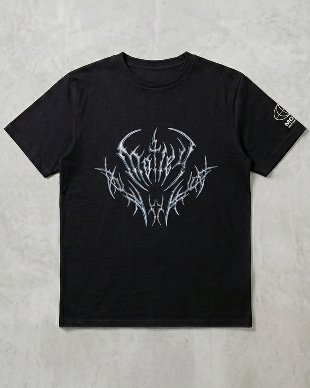Vista frontal plana de una camiseta negra de algodón sobre un fondo de cemento claro, con un diseño gráfico central estilo "heavy metal" en tonos plateados metálicos y un pequeño logo blanco en la manga izquierda.