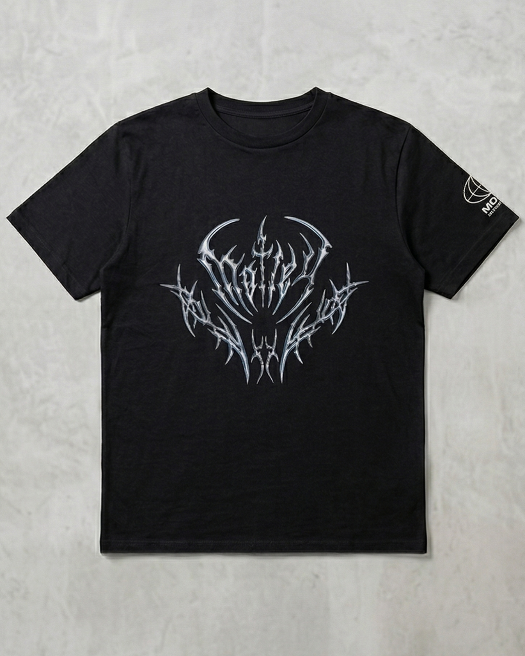 Vista frontal plana de una camiseta negra de algodón sobre un fondo de cemento claro, con un diseño gráfico central estilo "heavy metal" en tonos plateados metálicos y un pequeño logo blanco en la manga izquierda.