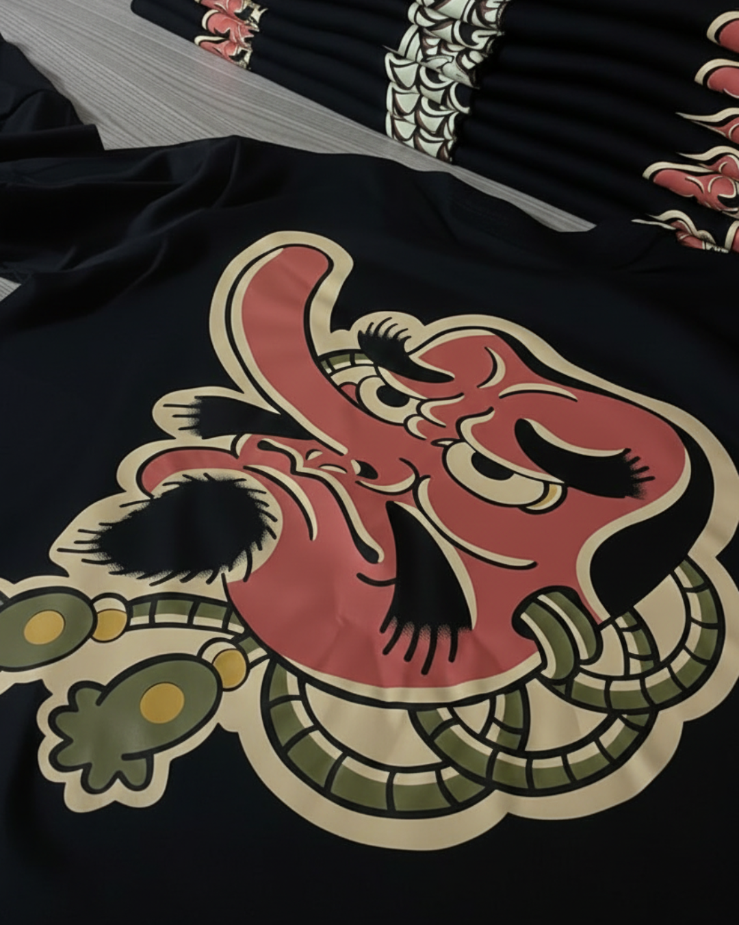 Vista trasera de la camiseta negra Wrath of Tengu de algodón exhibida sobre un fondo artístico japonés con olas y flores de cerezo. Se aprecia el gran diseño de la máscara Tengu de nariz larga y detalles verdes en la espalda