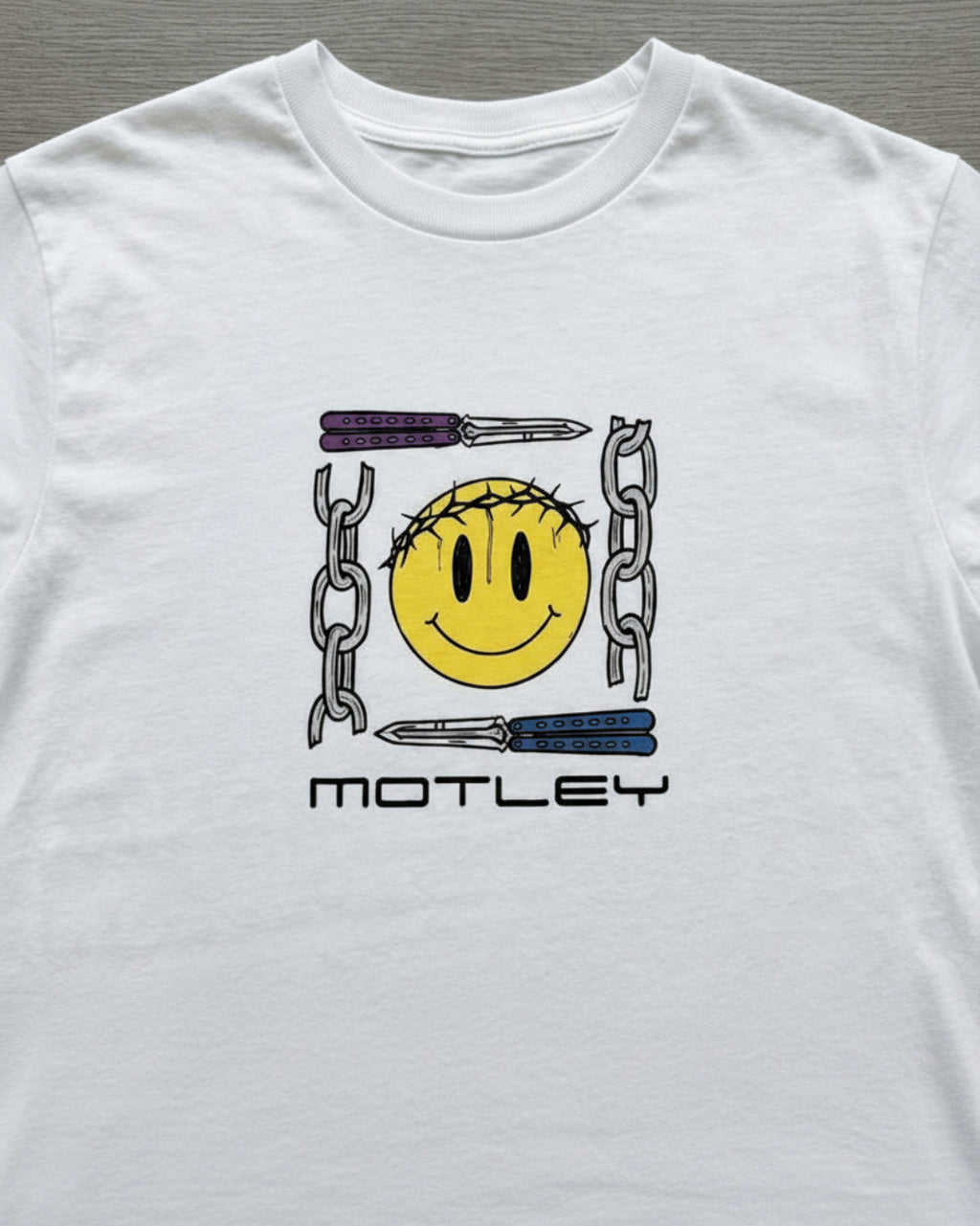 Detalle del estampado frontal de la camiseta Smiley Core de Motley. Ilustración de carita feliz con corona de espinas, navajas mariposa y cadenas. Calidad de impresión de autor en Tattoo Roots Colombia.
