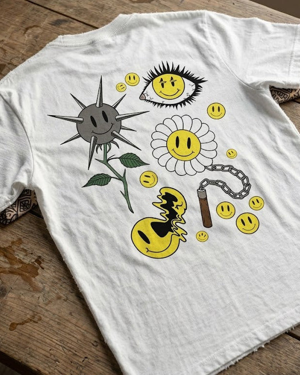 Diseño artístico en espalda de camiseta blanca Motley. Composición de ilustraciones tipo flash tattoo: smiley con espinas, ojo con pestañas de punta, margarita con nunchakus y smileys derretidos. Estética agresiva y artística de Tattoo Roots.