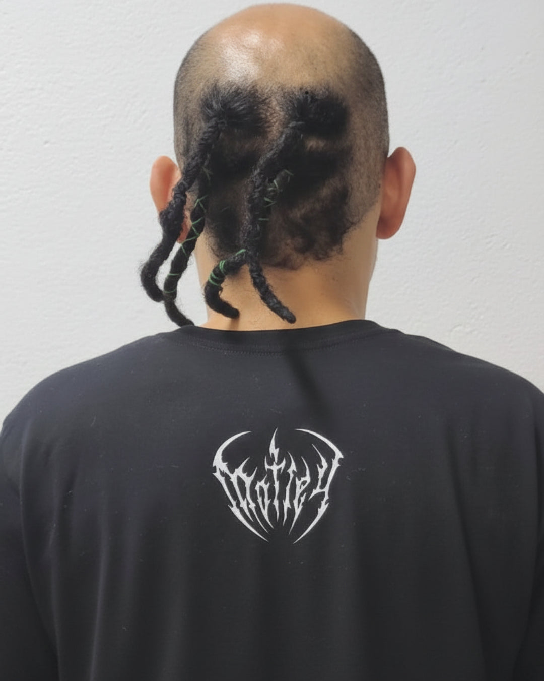 Modelo hombre de espalda mostrando el fit de la camiseta negra Motley. Detalle del logo gótico de autor en la nuca y trenzas estilo urbano. Streetwear oscuro de Tattoo Roots Colección Cruda Vibra.