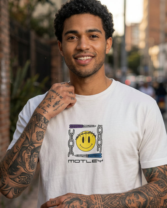 Modelo hombre posando de frente con camiseta blanca Motley Smiley Core. Se aprecia el ajuste de la prenda en el torso y el tamaño del estampado central. Streetwear colombiano Tattoo Roots.
