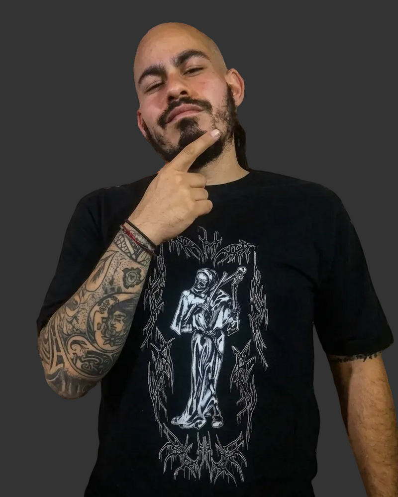 Modelo hombre con tatuajes en el brazo luciendo el frente de la camiseta negra Motley Tribal Reaper. Se aprecia el tamaño del estampado de la parca gótica en el pecho y el cuello redondo estructurado. Streetwear de autor Tattoo Roots.