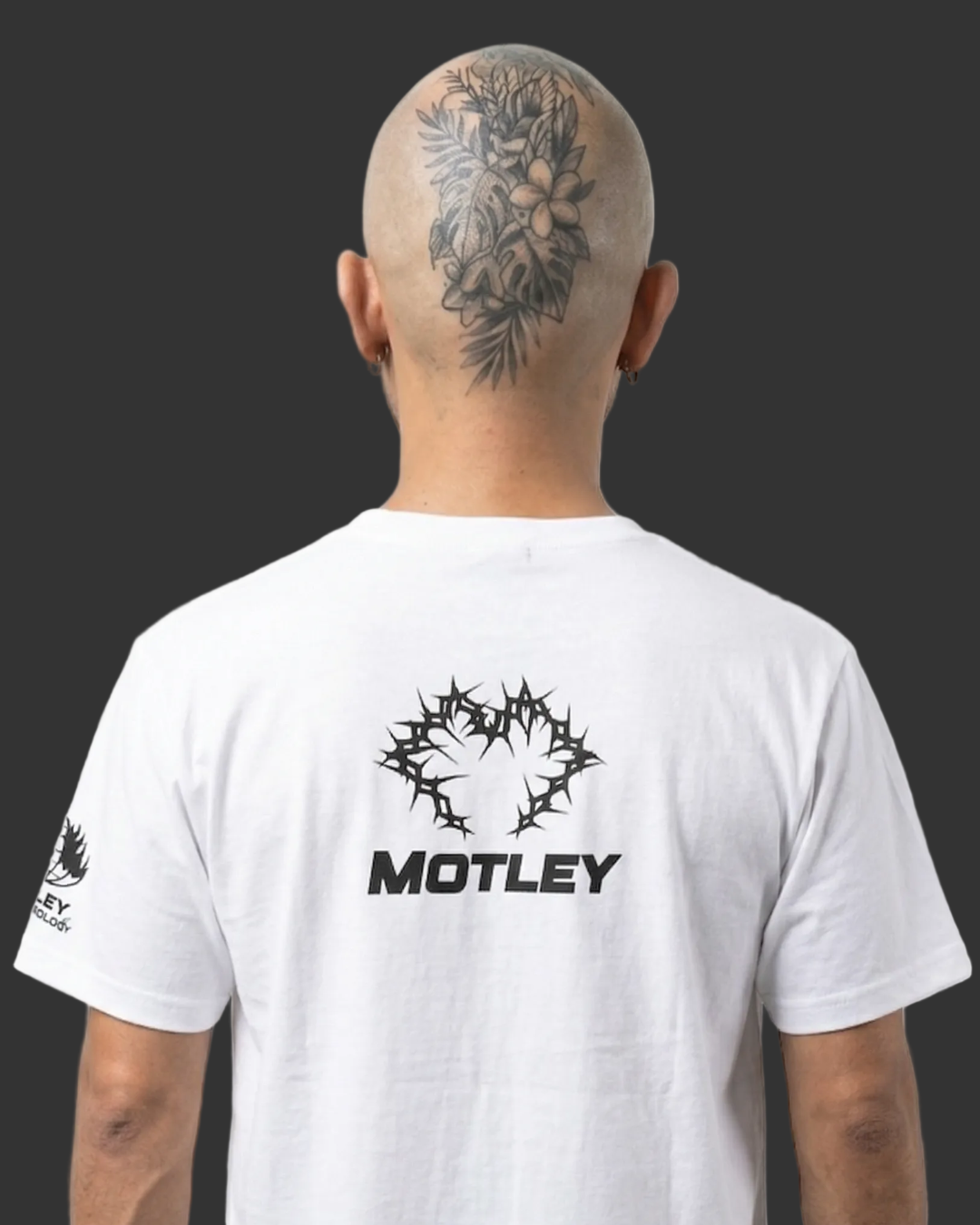 Vista de espalda de un modelo calvo con un gran tatuaje floral y de hojas en la parte posterior de la cabeza. Lleva una camiseta blanca con un gráfico central negro de espinas entrelazadas y la palabra "MOTLEY" debajo en fuente sans-serif negra, además de un gráfico negro más pequeño en la manga izquierda, contra un fondo blanco limpio.