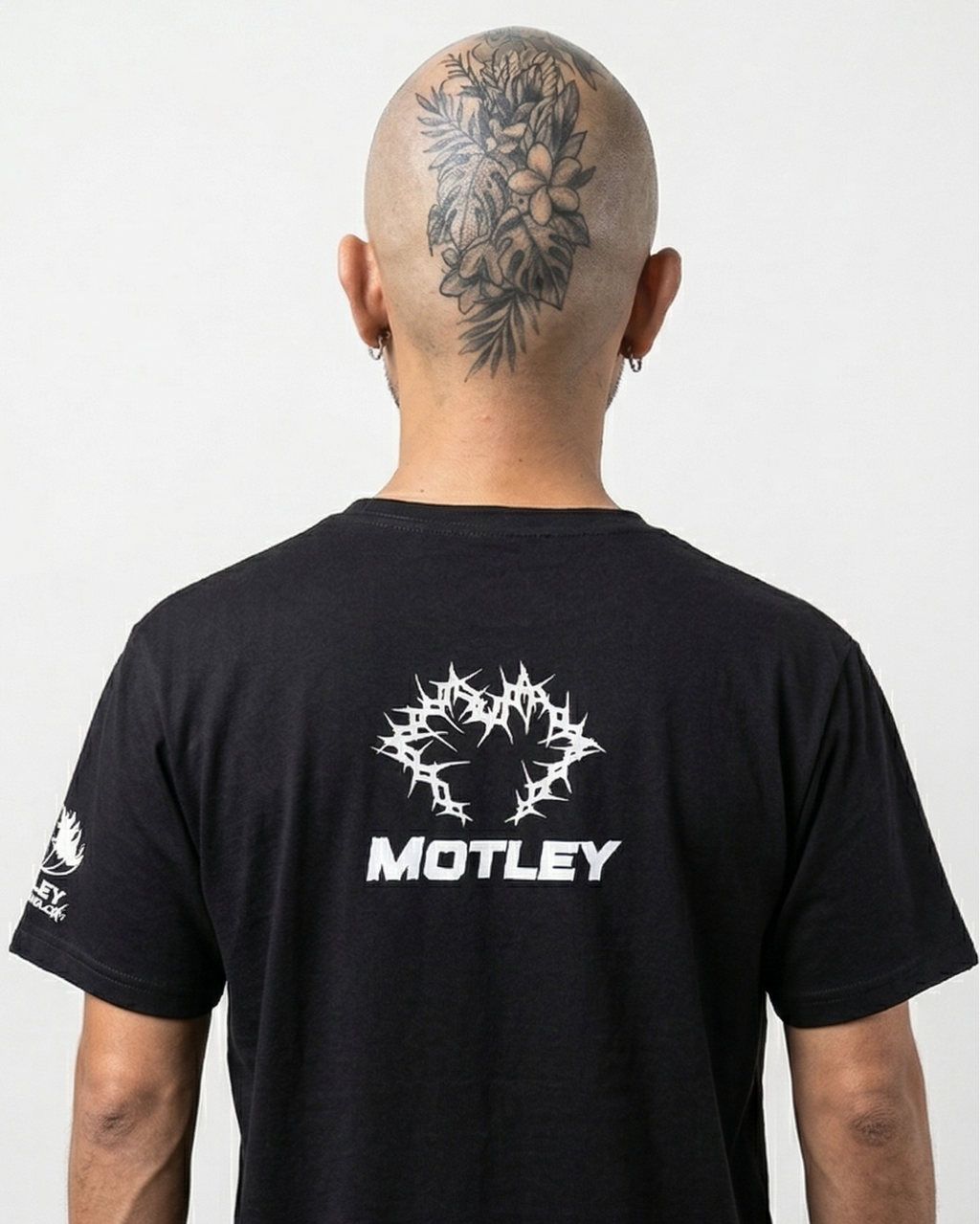 Vista de espalda de un modelo calvo con un gran tatuaje floral y de hojas en la parte posterior de la cabeza. Lleva una camiseta negra con un gráfico central blanco de espinas entrelazadas y la palabra "MOTLEY" debajo en fuente sans-serif blanca, además de un gráfico blanco más pequeño en la manga izquierda, contra un fondo negro limpio.