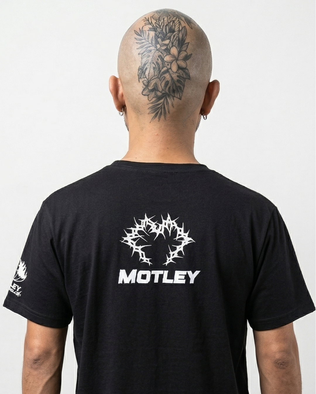 Vista de espalda de un modelo calvo con un gran tatuaje floral y de hojas en la parte posterior de la cabeza. Lleva una camiseta negra con un gráfico central blanco de espinas entrelazadas y la palabra "MOTLEY" debajo en fuente sans-serif blanca, además de un gráfico blanco más pequeño en la manga izquierda, contra un fondo negro limpio.