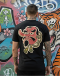 Vista de la espalda de un modelo usando la camiseta negra con el diseño artístico de la máscara Tengu de nariz larga. Se aprecia el detalle del estampado de alta calidad y el fondo urbano con mural de tigre.