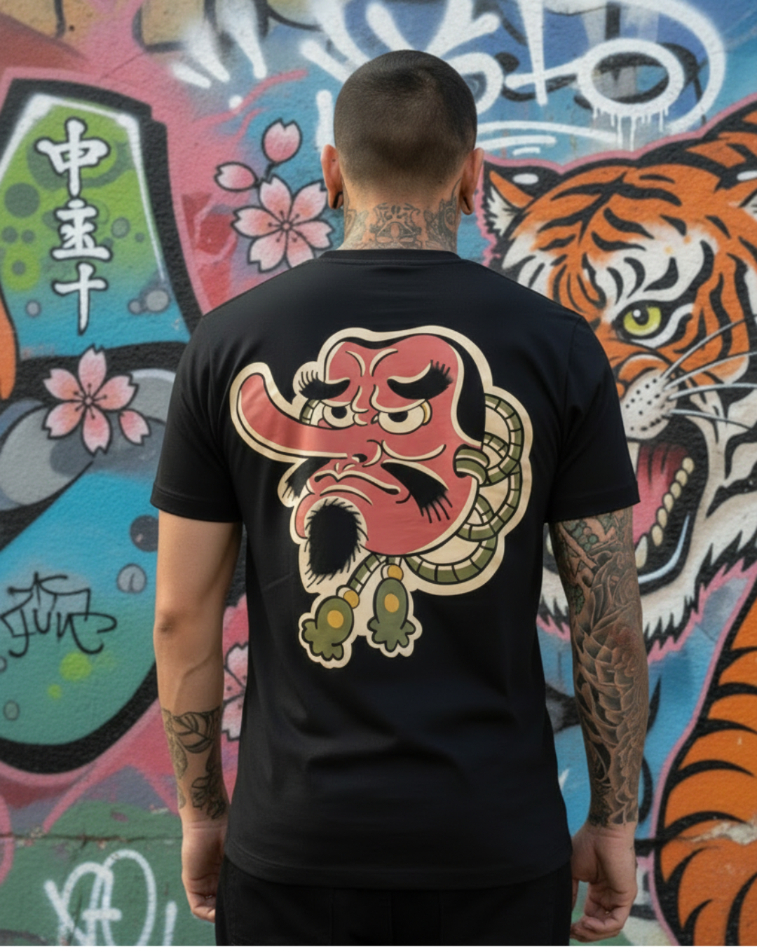 Vista de la espalda de un modelo usando la camiseta negra con el diseño artístico de la máscara Tengu de nariz larga. Se aprecia el detalle del estampado de alta calidad y el fondo urbano con mural de tigre.
