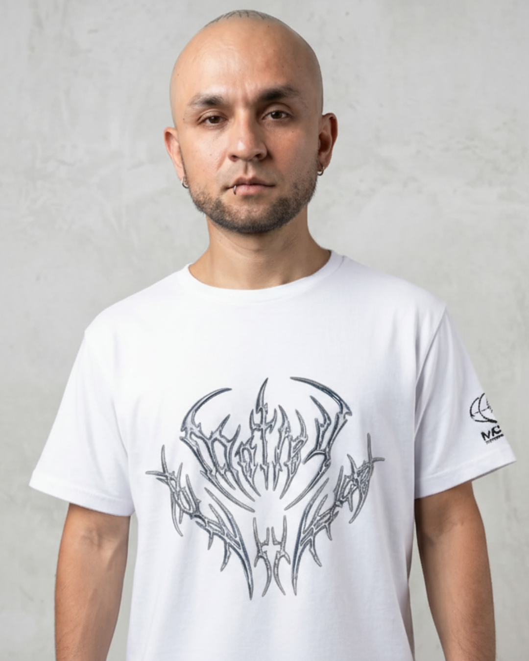 Un modelo masculino calvo con una camiseta blanca de manga corta, con un gran diseño gráfico metálico plateado y azul de estilo tribal y de heavy metal en el pecho, de pie contra una pared de concreto texturizada.