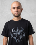 Un modelo masculino calvo con una camiseta negra de manga corta, con un gran diseño gráfico metálico plateado y azul de estilo tribal y de heavy metal en el pecho, de pie contra una pared de concreto texturizada.