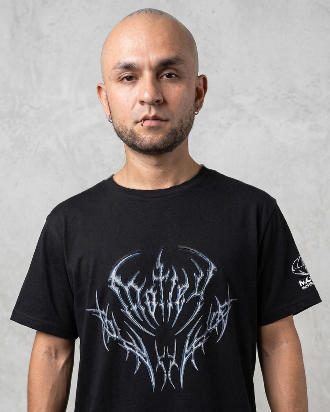 Un modelo masculino calvo con una camiseta negra de manga corta, con un gran diseño gráfico metálico plateado y azul de estilo tribal y de heavy metal en el pecho, de pie contra una pared de concreto texturizada.