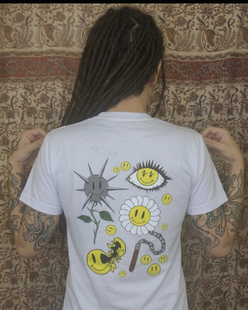Mujer con rastas y brazos tatuados posando de espalda con la camiseta blanca Motley. Se aprecia el fit y el tamaño real del diseño Smiley Core de autor. Streetwear colombiano en Tattoo Roots.